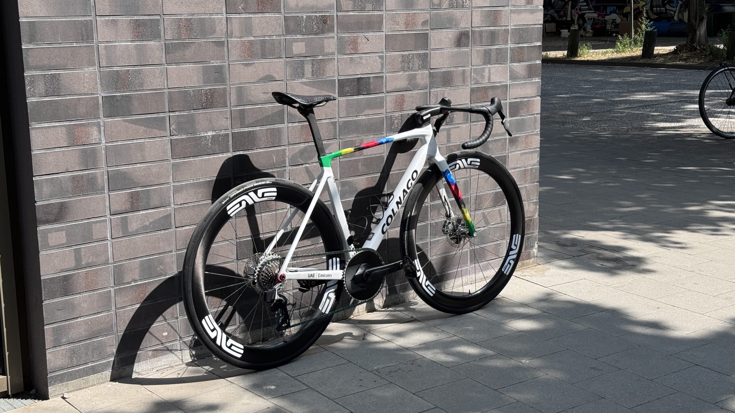 Weißes Colnago-Rennrad mit farbigen Akzenten, ENVE-Laufrädern und SRAM-RED-Antrieb vor einer Backsteinwand